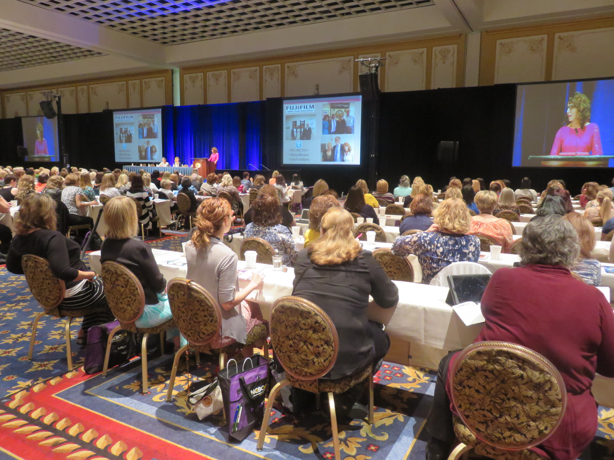Conference attendees at round tables watching Dr. Cappello's keynote on large screens