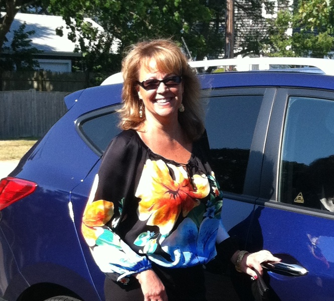 Suellen in a floral top, standing by a blue car