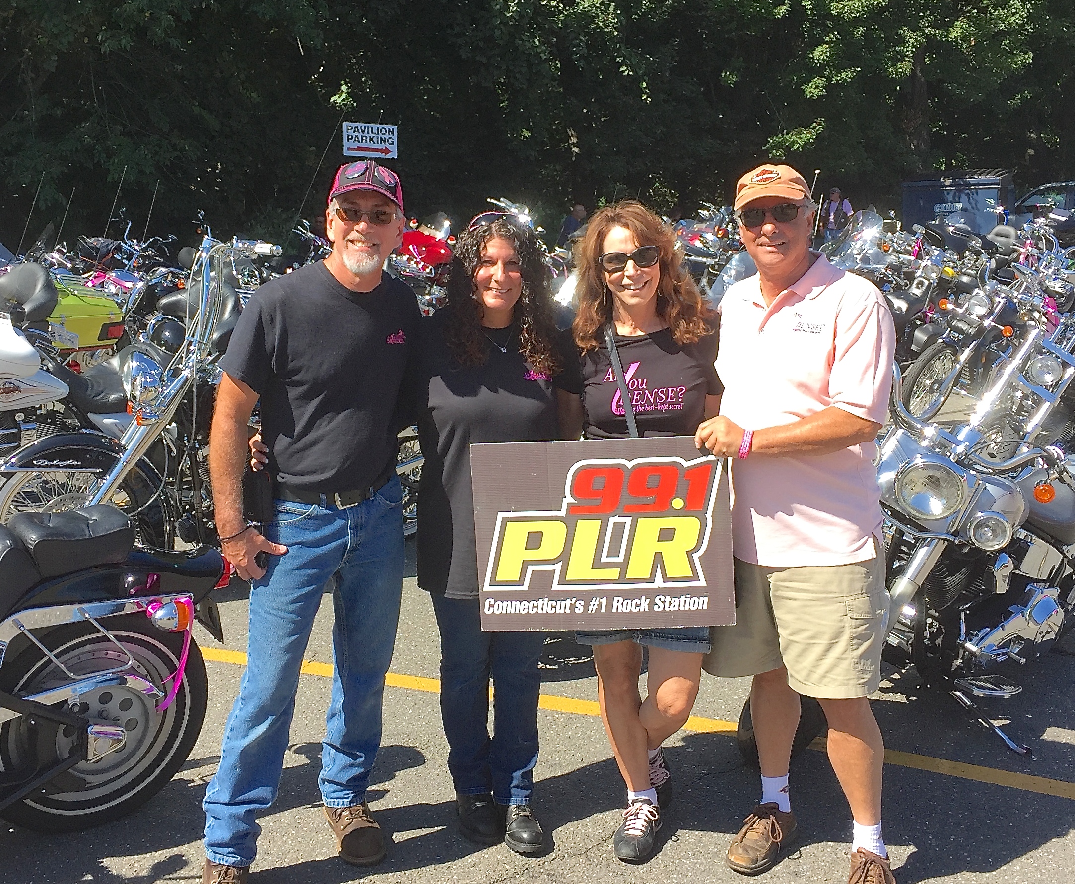 Four Ribbon Ride organizers holding a 99.1 PLR sign in front of rows of parked motorcycles