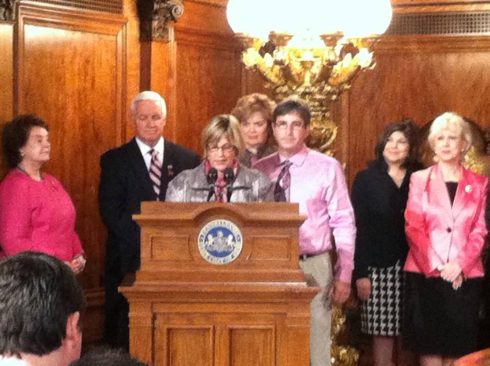 Cindy Spinello speaking at a podium, flanked by her husband Greg and supporters