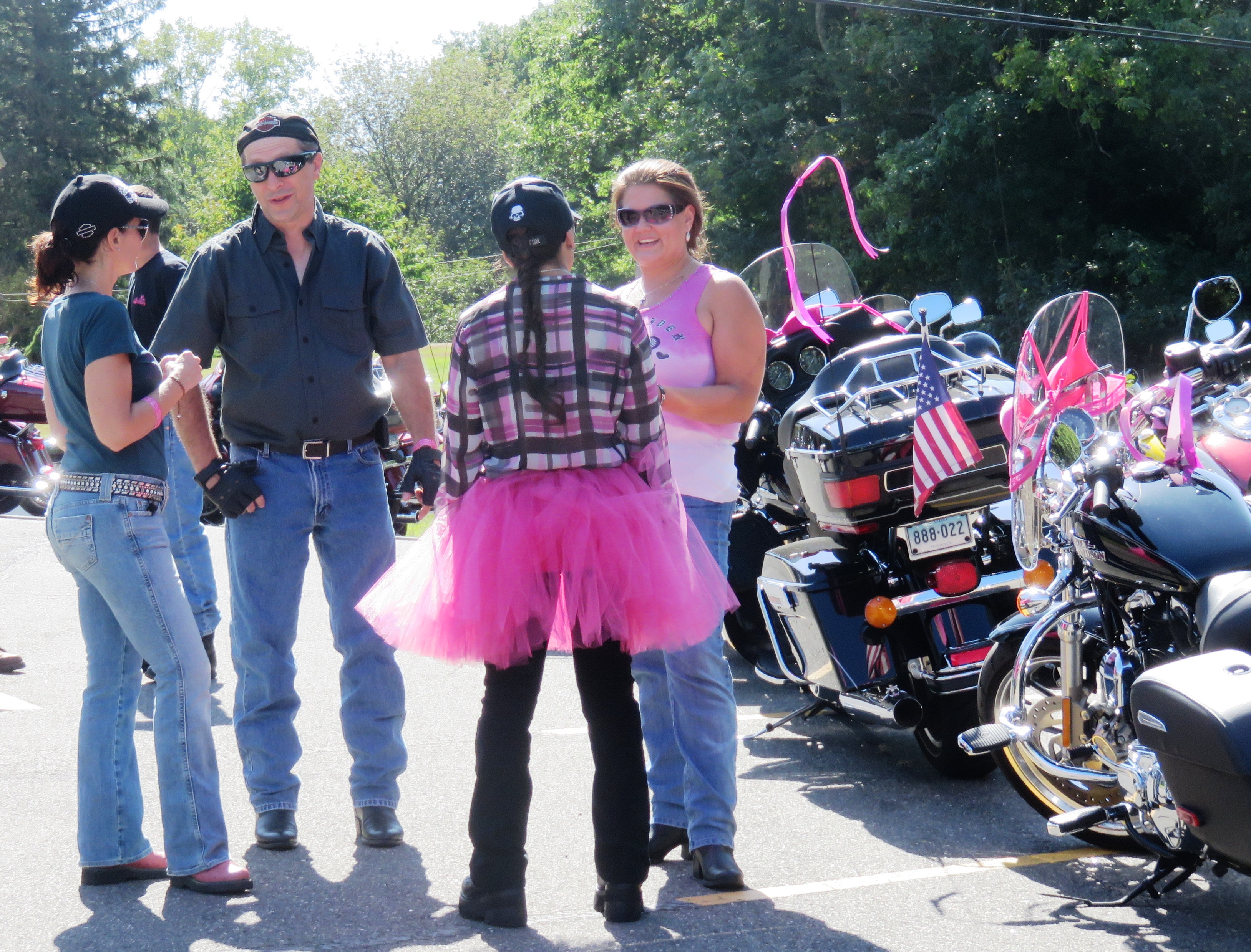 Riders, one in a pink tutu, chatting beside motorcycles decorated with pink ribbons